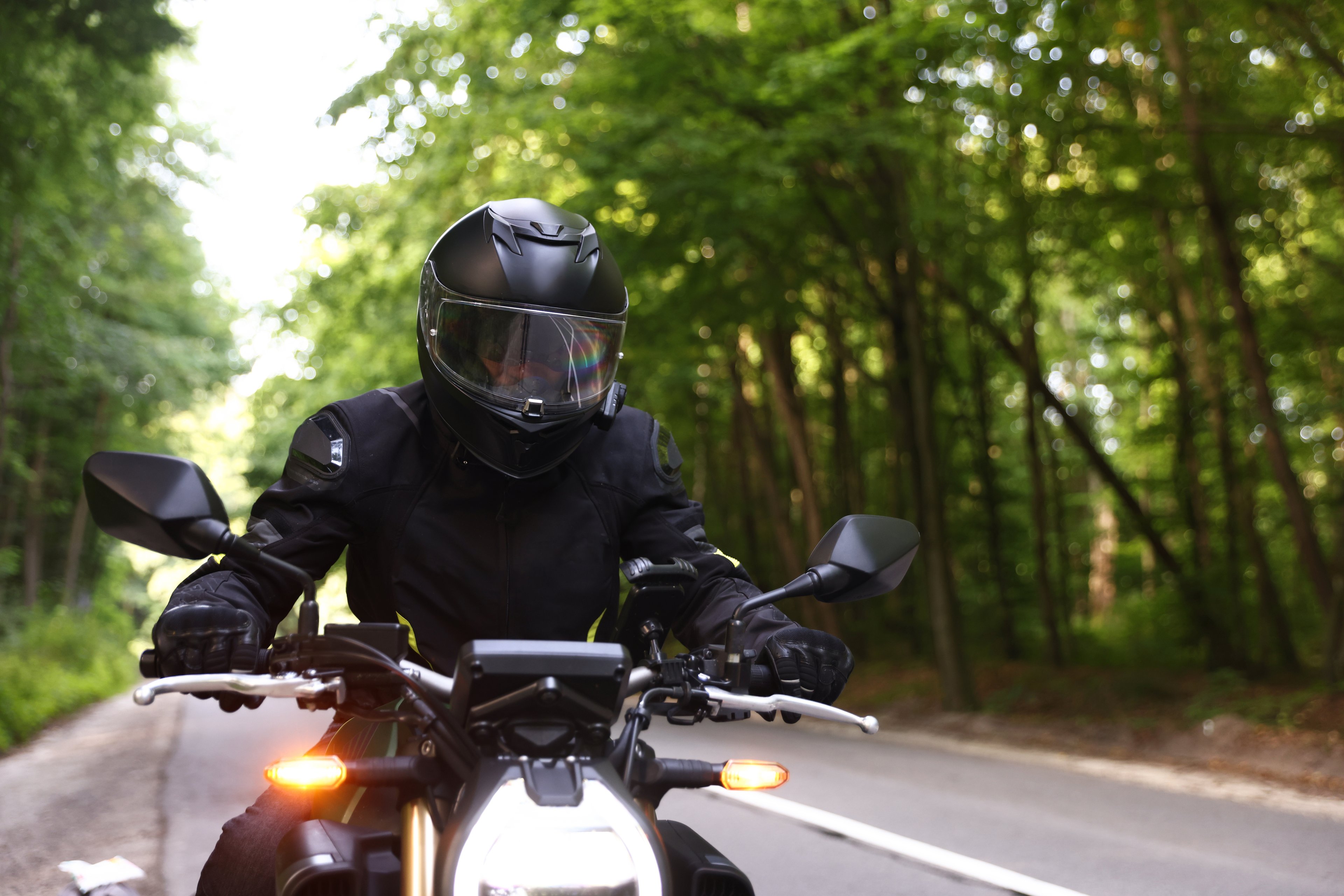 A motorcyclist in a black helmet and riding jacket on a tree-lined two-lane road